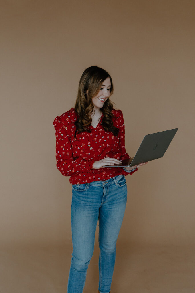 Woman in red shirt and blue jeans against a brown background holding and looking down at gray MacBook.